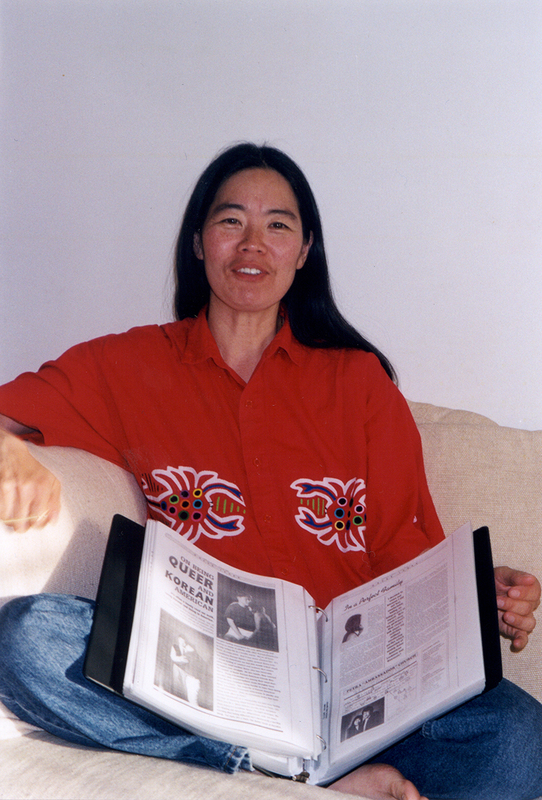 Alison Kim, a person with long black hair and a red shirt with stylized crabs on it, sits on a couch with one leg up and a binder scrap book on her lap.